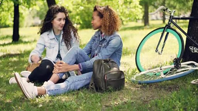非裔美国女孩正在和她的白人朋友坐在公园的草地上聊天，朋友们在聊天和微笑。现代自行车和背包是可见的。