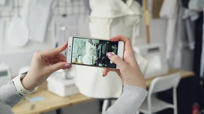 妇女手持智能手机并拍摄剪裁假人的特写镜头，并将半成品服装固定在上面。小企业概念中的现代技术。