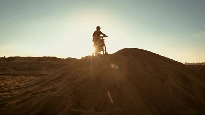 专业的摩托车越野者骑着FMX摩托车在沙丘上行驶，在沙丘上停下来欣赏美丽的日落景色。
