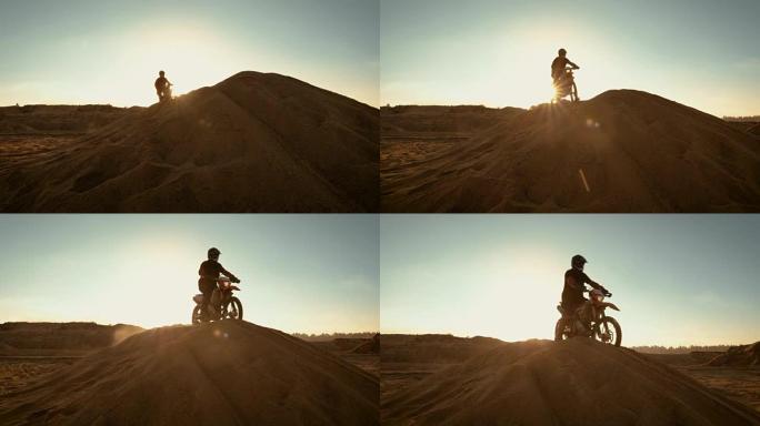 专业的摩托车越野者骑着FMX摩托车在沙丘上行驶，在沙丘上停下来欣赏美丽的日落景色。