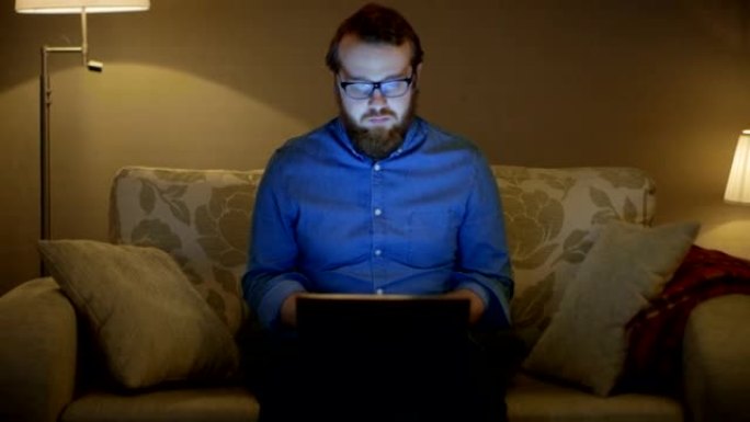 一个男人坐在客厅的沙发上，膝盖上有笔记本电脑的肖像。他在工作。落地灯打开。