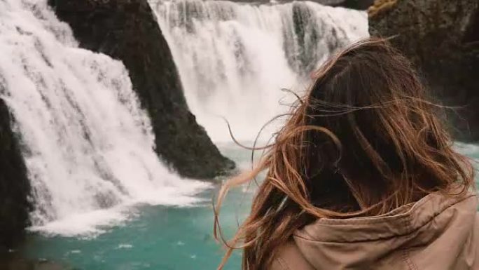 年轻有魅力的女人独自站着，看着冰岛强大的瀑布，风吹拂的特写镜头