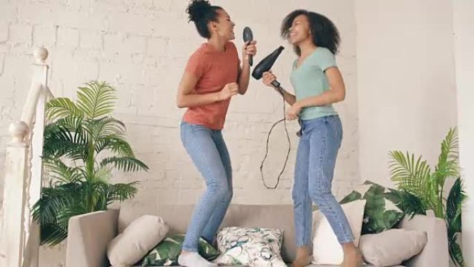 混合种族年轻有趣的女孩在沙发上用吹风机和梳子跳舞唱歌。姐妹们在客厅享受休闲的乐趣