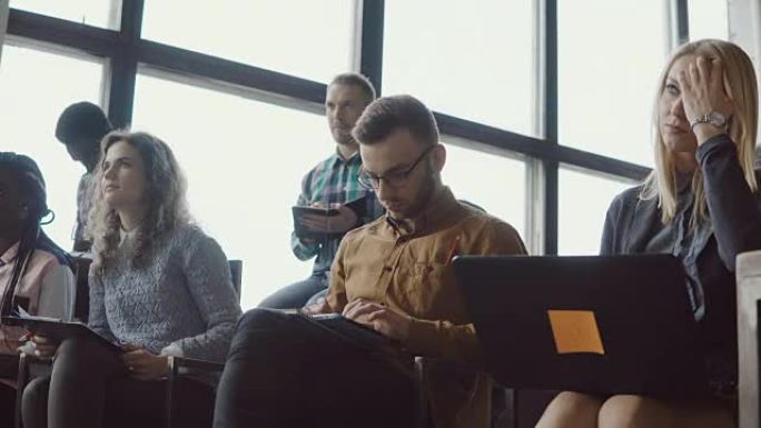 一群年轻人坐在阁楼办公室听研讨会。使用笔记本电脑和平板电脑的学生做笔记