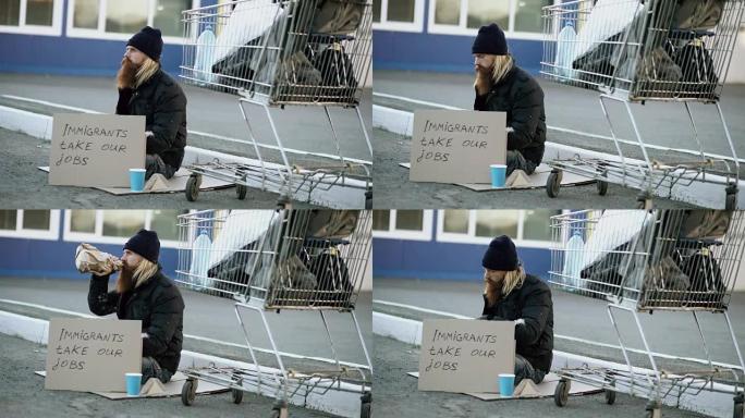 大胡子的年轻无家可归的人，拿着纸板坐在购物车附近，在寒冷的日子里喝酒。欧洲的移民危机