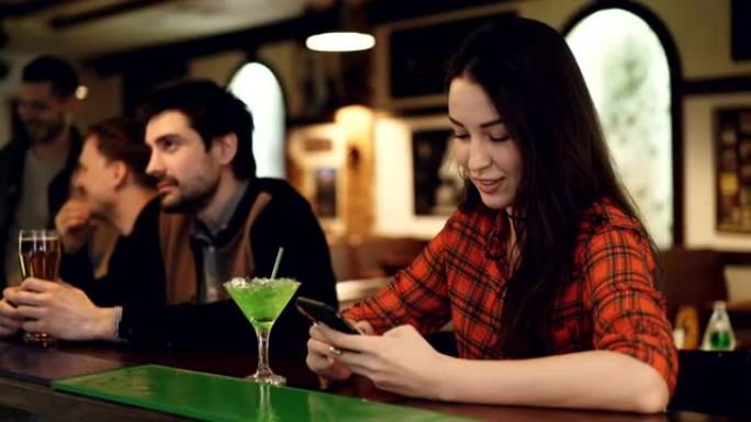 年轻的女学生正在使用智能手机，坐在高档酒吧里喝鸡尾酒。她正在触摸屏幕并微笑。现代沟通方式概念。