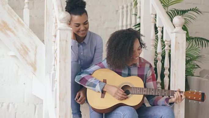 两个巴西卷发女孩姐妹坐在楼梯上练习弹吉他。朋友在家里玩得开心，唱歌