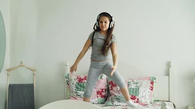 有趣可爱的小女孩戴着无线耳机跳舞，在假期早上在舒适的轻便卧室里跳到家里的床上玩得开心