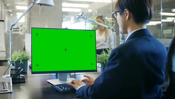 在办公室里，商人在办公桌前工作，使用带有绿屏模型的个人计算机。同事进入办公室，在她的办公桌前进行。