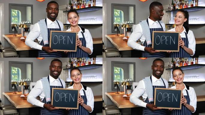 微笑的服务员和女服务员在餐厅拿着石板，上面挂着开放的牌子