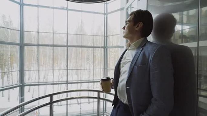 年轻英俊的商人穿着西装喝咖啡，在现代商务中心的eleavator里往下走