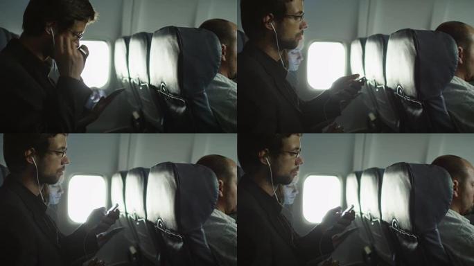 年轻人正在飞机上听音乐，一个女人正在窗户旁边的背景中使用平板电脑。