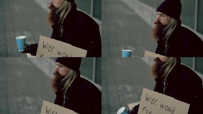 无家可归的年轻人的特写镜头乞求摇钱杯注意人们在城市人行道上的乞g附近行走