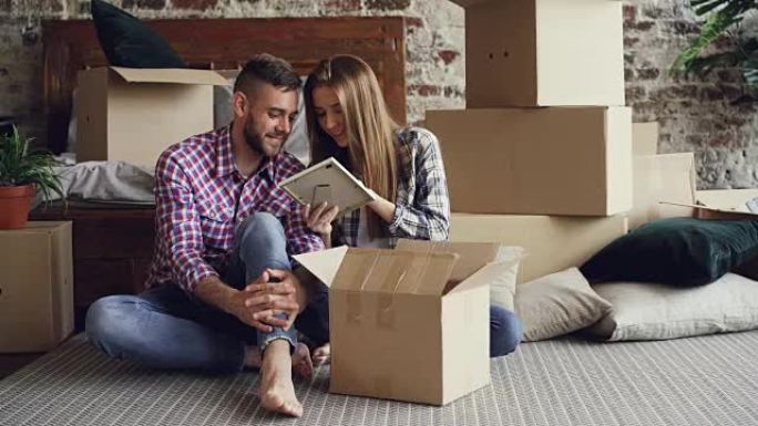 已婚夫妇坐在新房子的地板上时正在打开私人物品的包装。年轻人有说有笑，纸箱和双人床可见。