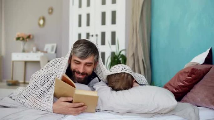 有爱心的父亲和可爱的儿子正在读可怕的故事，躺在家里的床上躲在毯子下。快乐的童年，为人父母和爱的观念。