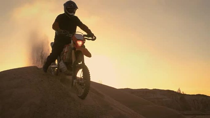 专业的摩托车越野赛摩托车骑手在沙丘上驾驶，并在顶部停下。现在是日落时分，跑道上布满了烟雾。