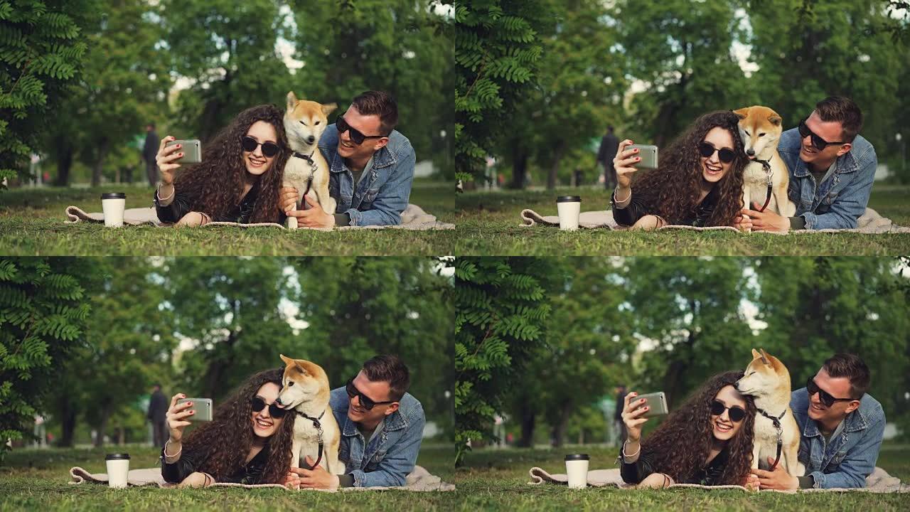 年轻女子正在公园里与男友和宠物狗躺在草地上自拍，可爱的动物在打喷嚏和舔鼻子，人们在笑。