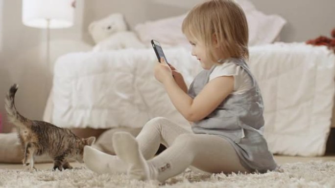 可爱的小女孩坐在地板上，用智能手机拍摄她的条纹小猫四处走动的视频。慢动作。