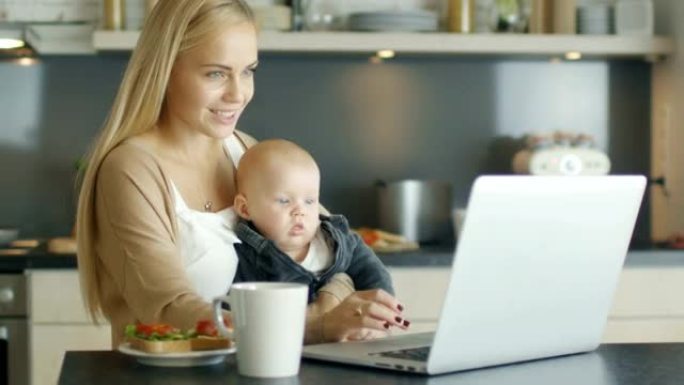在厨房里，美丽的微笑母亲在膝盖上抱着可爱的孩子时使用笔记本电脑。
