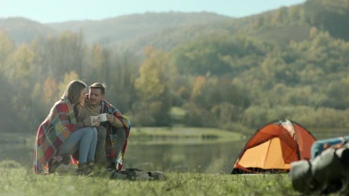 男人和女人坐在背上的毯子上，在湖边露营时喝咖啡