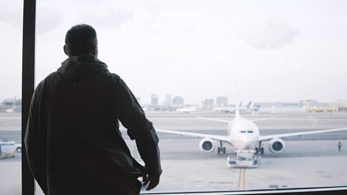 男性旅行者肩上背着大包，走到机场候机室候机楼的窗户，欣赏飞机和汽车的景色