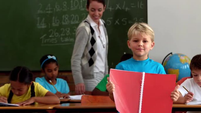 教室里拿着红色记事本的小男孩对着相机微笑