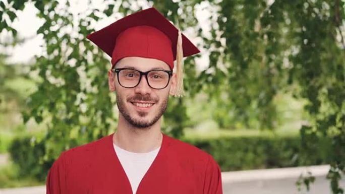 戴着眼镜，毕业礼服和灰浆板微笑着看着站在户外的相机的大胡子青年毕业学生的肖像。人和教育理念。