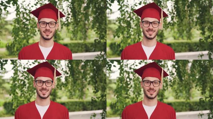 戴着眼镜，毕业礼服和灰浆板微笑着看着站在户外的相机的大胡子青年毕业学生的肖像。人和教育理念。