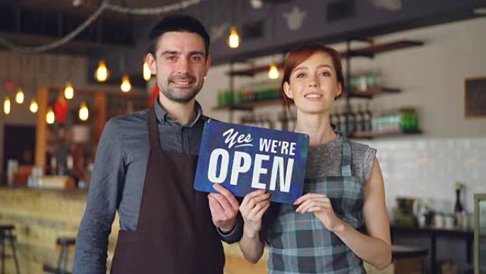 快乐迷人的人围着围裙的咖啡馆工作人员站在咖啡店里，举着 “是的，我们是开放的” 标志，微笑着。开启新