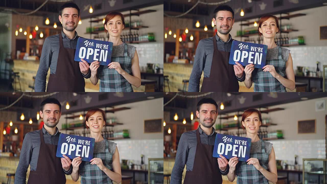 快乐迷人的人围着围裙的咖啡馆工作人员站在咖啡店里，举着 “是的，我们是开放的” 标志，微笑着。开启新