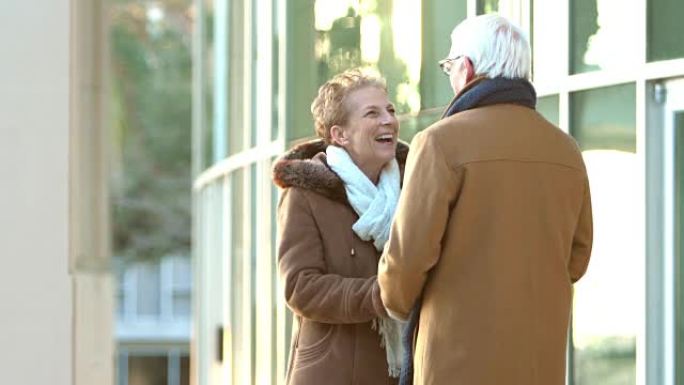 穿着大衣的老年情侣在大楼外聊天，拥抱
