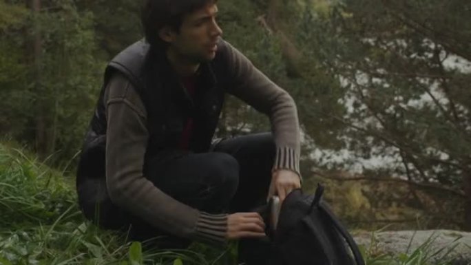 男人坐在森林山顶上，从包里拿出热水瓶。