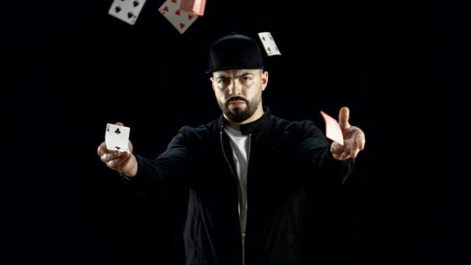 专业的街头魔术师在一个黑暗的衣服和一个帽子发射两个喷气的纸牌到空中在慢动作。背景是黑色的。
