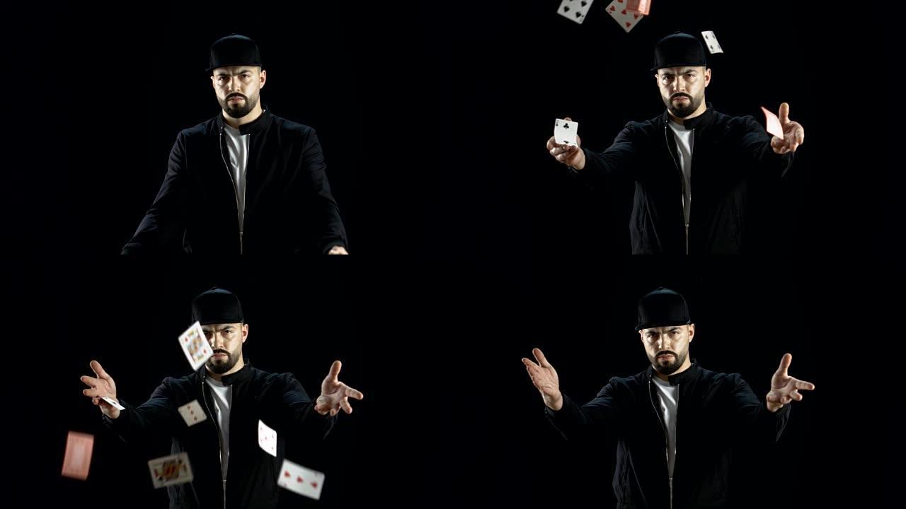 专业的街头魔术师在一个黑暗的衣服和一个帽子发射两个喷气的纸牌到空中在慢动作。背景是黑色的。
