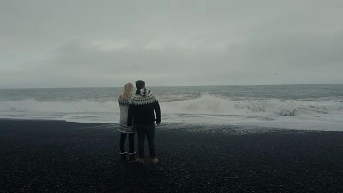 年轻夫妇站在巨魔脚趾岩石附近的黑色火山海滩上，在冰岛看波浪的鸟瞰图