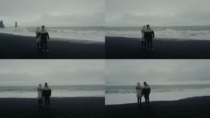 年轻夫妇站在巨魔脚趾岩石附近的黑色火山海滩上，在冰岛看波浪的鸟瞰图