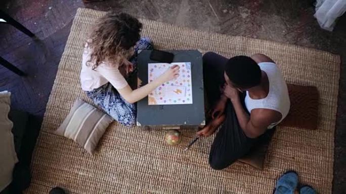 多种族夫妇坐在地板上玩棋盘游戏的俯视图。女人掷骰子，拿卡。男人等待转弯