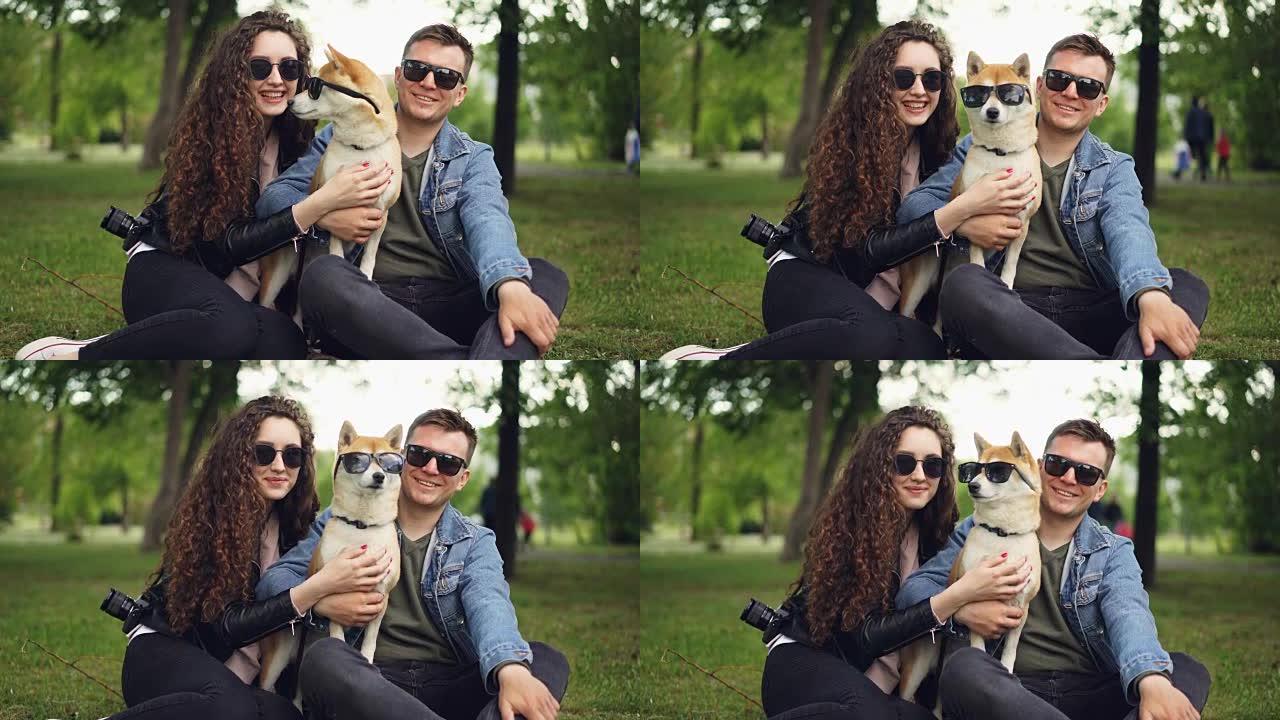 可爱的情侣和柴犬的肖像都戴着墨镜看着相机，坐在公园的草地上微笑。现代生活方式，可爱的动物和自然概念。