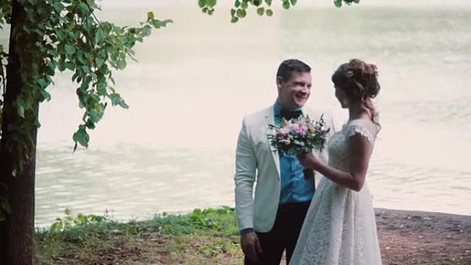 相爱的幸福夫妇分享他们的婚礼。他们站在河岸上，有说有笑。漂亮的服装，花束