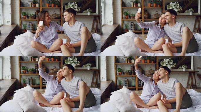 有趣的年轻人的男朋友和女朋友正坐在家里的床上和愚蠢的脸一起自拍，玩得开心，接吻。自画像和重新定义概念