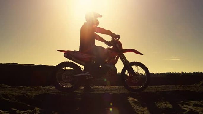 侧面镜头的专业摩托车越野赛摩托车骑手坐在他的FMX土地车准备驾驶越野赛道。它发生在采石场和美丽的日落