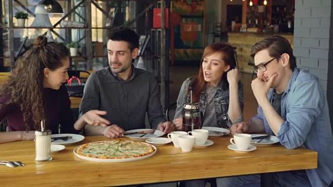开朗的年轻朋友坐在现代咖啡馆的餐桌旁聊天和打手势分享新闻。可以看到大披萨，杯子和盘子，桌子和椅子。