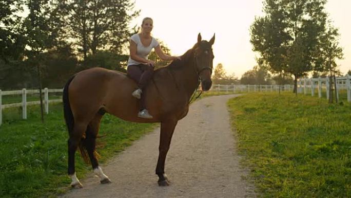 慢动作女孩坐在一匹马上，双腿交叉，在日出时抚摸他
