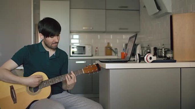 开朗的年轻人坐在厨房里学习在家中使用笔记本电脑弹吉他