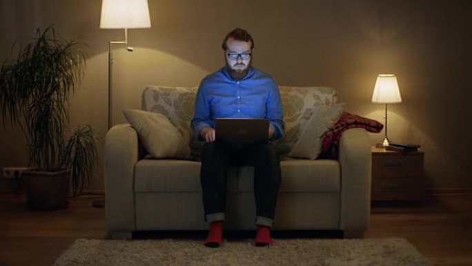 一个男人坐在客厅的沙发上，膝盖上有笔记本电脑的肖像。他在工作。他直视镜头。落地灯打开。