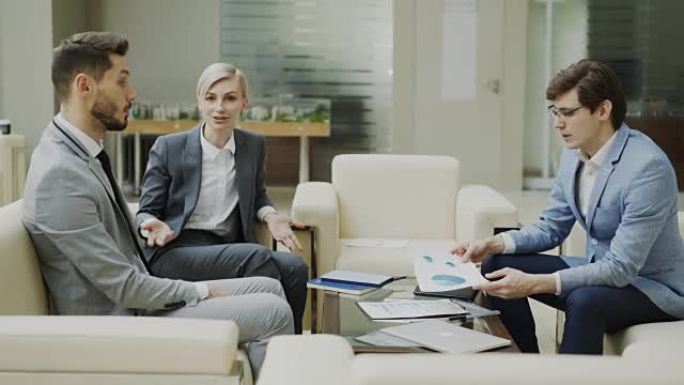 商人与坐在现代办公厅沙发上的男女商业伙伴讨论财务报告的平移镜头