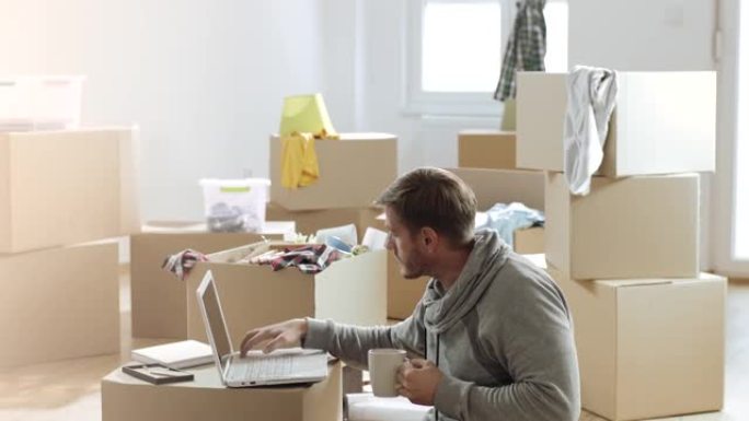 在装满未包装盒子的新公寓里工作的人在笔记本电脑上喝咖啡