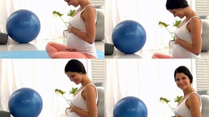 微笑的孕妇坐在垫子上抚摸她的肚子