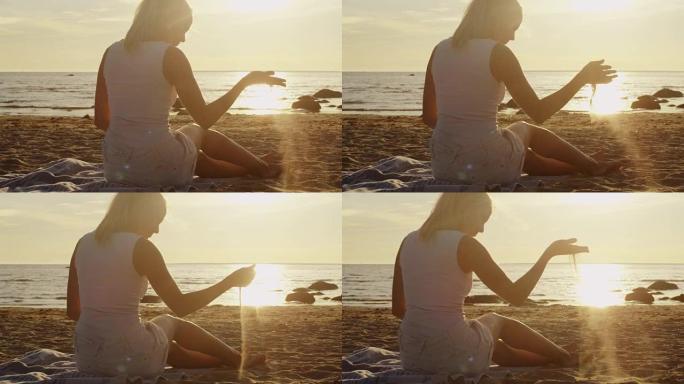 穿着白布的年轻女孩坐在沙滩上，在日落的灯光下玩沙子