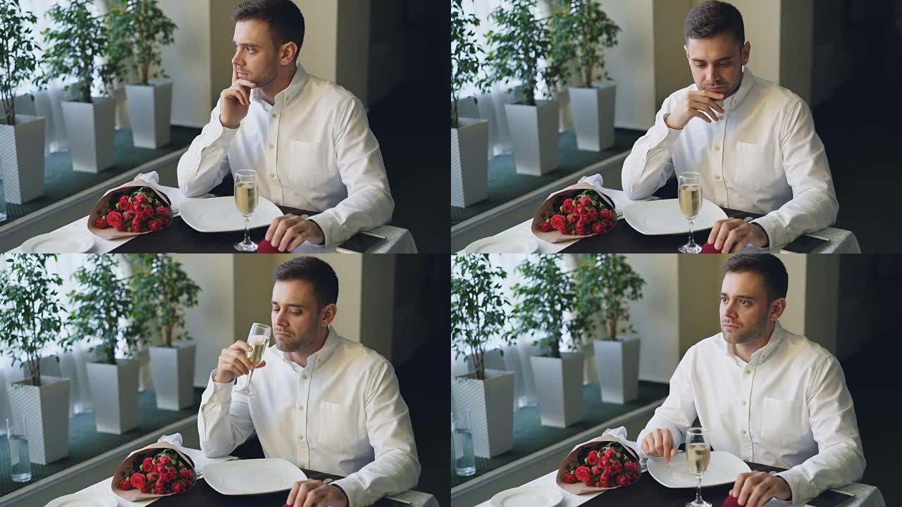 紧张的男人独自一人坐在餐厅的餐桌旁，喝着香槟，等待女友，然后离开。一束红玫瑰，珠宝盒和智能手机可见。